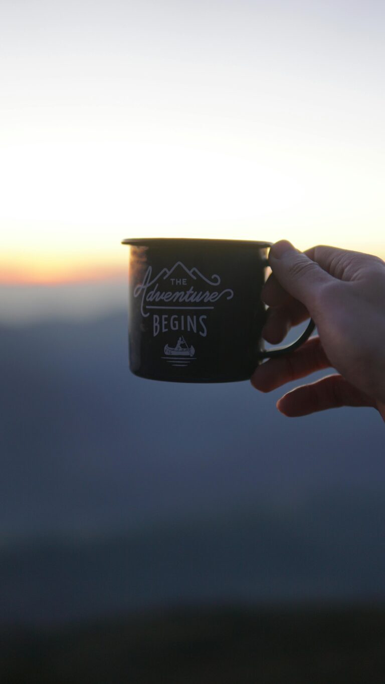 Silhouette of hand holding a mug reading 'The Adventure Begins' during twilight outdoors.